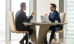 Two businessmen in suits sitting at a desk speaking.