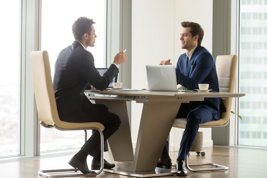 Two businessmen in suits sitting at a desk speaking.